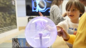 A young boy smiling enthusiastically as he touches a glowing purple plasma ball exhibit at the UCLA Exploring Your Universe science event.