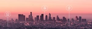 A pink and orange twilight sky silhouettes the Downtown Los Angeles skyline. Three white circular icons overlay the scene, showing a bus, a dollar-sign coin, and a walking figure. White lines extend downward from each circle toward the cityscape.
