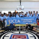 A large group of UCLA student volunteers poses together inside an indoor basketball court, standing and reclining behind a large blue and gold banner that reads "UCLA COMMUNITY PARTNER."