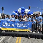 A large group of UCLA student volunteers and Los Angeles Mayor Karen Bass smile together on a street parade float behind a blue and gold banner reading "UCLA COMMUNITY PARTNER." An arch of blue and white balloons decorates the background under a clear sky.