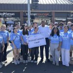 A group of UCLA student volunteers wearing light blue shirts poses on a city street alongside a man in a white button-down shirt. Two volunteers in the front hold a white sign that reads "Brentwood Community Council."