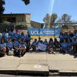 A large group of UCLA student volunteers poses together outdoors on a sunny sidewalk in front of a chain-link fence at Fairburn Ave. Elementary School.