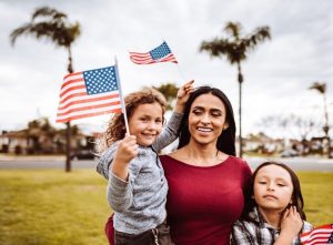 A happy family of three celebrating an American holiday outdoors, with a young child holding up a small US flag against a background of palm trees.