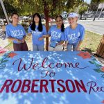 Four young women wearing light blue UCLA Volunteer shirts stand outdoors behind a freshly painted, bright blue sign that reads "Welcome To ROBERTSON Rec."