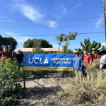 A group of community volunteers poses outdoors behind low garden shrubs at the Good Earth Community Garden. They stand on either side of a large blue and gold banner that reads "UCLA COMMUNITY PARTNER." Green trees, a wooden utility pole, and a clear blue sky are visible in the background.