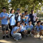A group of UCLA volunteers poses together on a sunny sidewalk, with some crouching and holding potted plants while making hand signs.