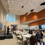 A speaker stands on a stage addressing a large crowd seated at round tables during an event inside a spacious, modern hall. Digital presentation screens line the left wall under high ceilings.