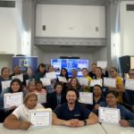 A large group of smiling event participants pose together indoors while proudly holding up printed certificates of completion. A large television screen displays slides in the background.