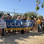 A large group of volunteers poses together on a dirt path in front of a light rail station platform under a clear blue sky. They are holding a large blue and gold banner that reads "UCLA COMMUNITY PARTNER" alongside a small white sign in the foreground reading "Westwood Greenway." Green trees and overhead power lines frame the background of the sunny scene.