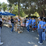 A group of UCLA student volunteers wearing light blue shirts gathers outdoors in a paved parking lot area to receive instructions before a community service event.
